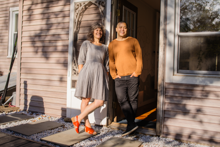 A man in an orange sweater and a woman in a gray dress stand in the doorway of a house smiling and leaning into one another.