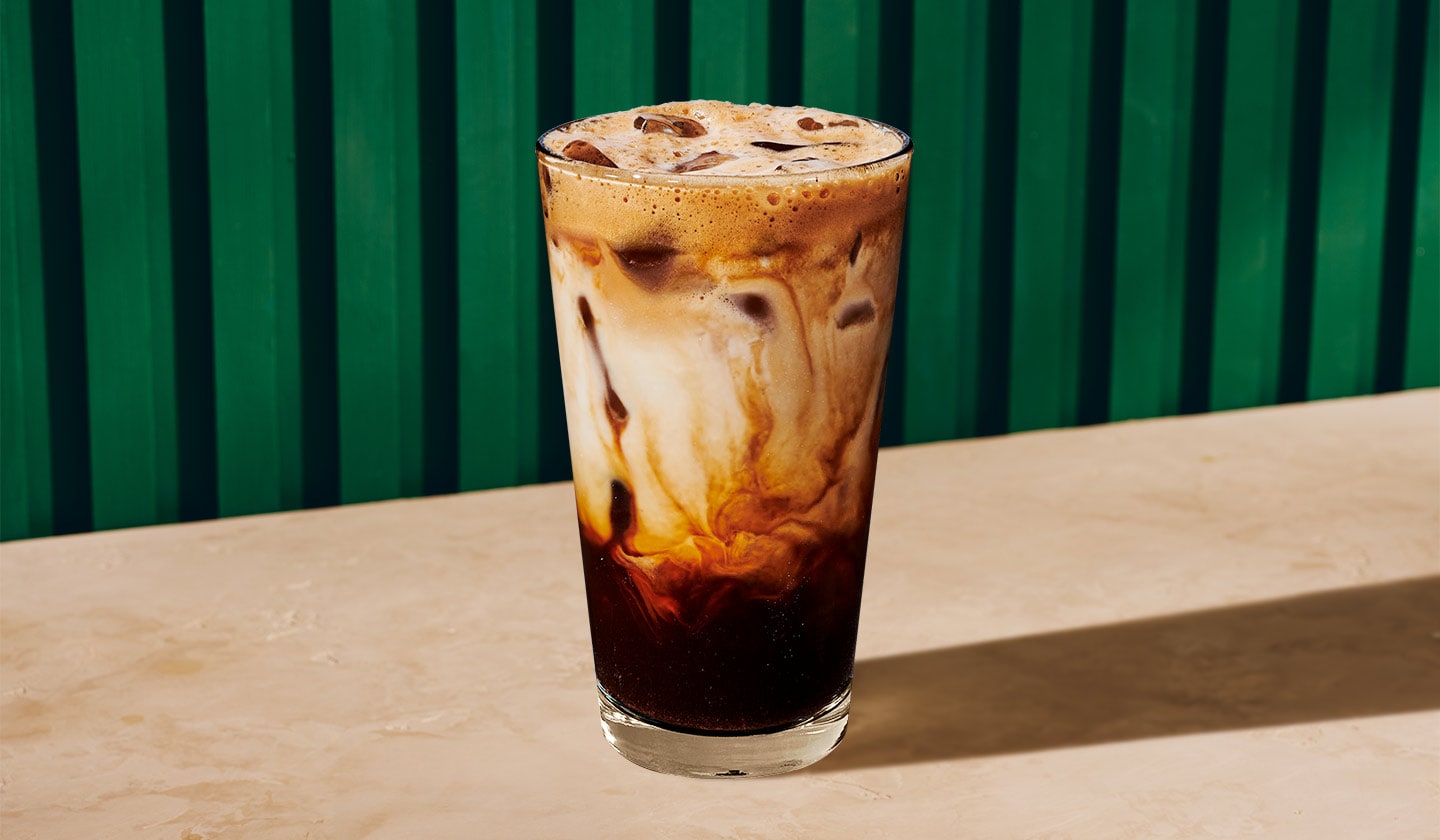 An Iced Brown Sugar Oatmilk Shaken Espresso sitting on a table top in front of a green striped background.
