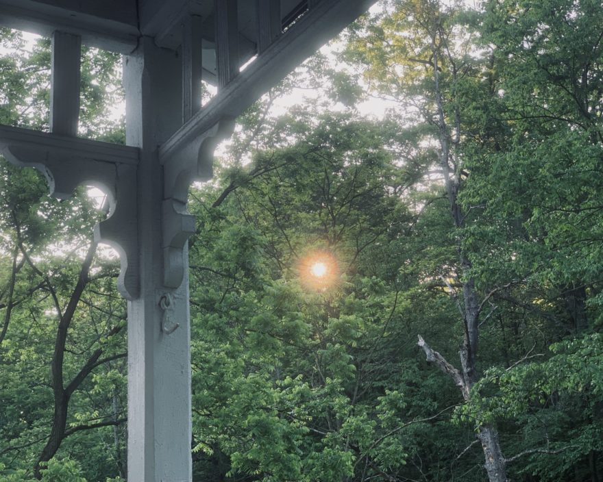view of the sun rising through green trees from a porch balustrade