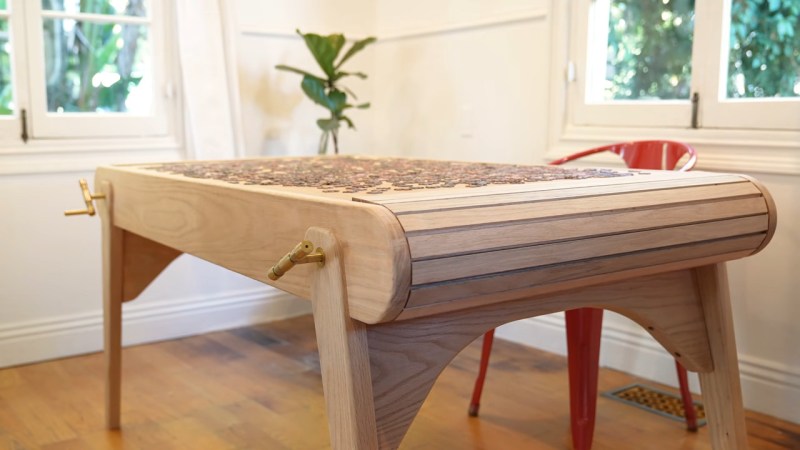 A wooden table with a puzzle on top of it sits in an off white room with a light wood floor. A red chair sits behind the table and the slats of the rolled away tambour top are visible.