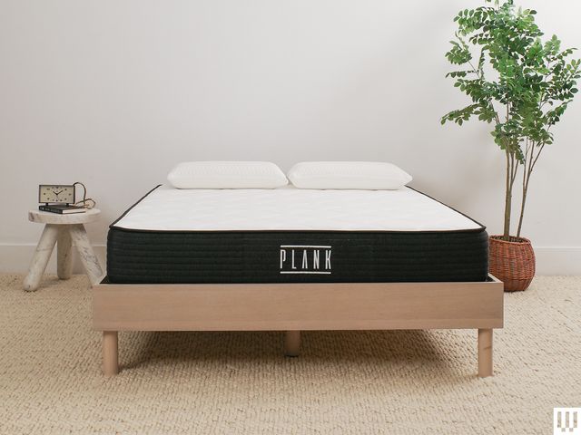Plank Firm Mattress a white mattress with blue trim sitting on a minimalist wooden frame with a nightstand and potted...