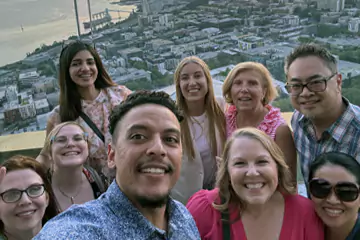 A group selfie of smiling coworkers with a cityscape in the background, taken from a high vantage point at sunset.